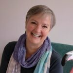 A middle aged white woman with short hair wrapped in a scarf laughs next to a wooden statue of a flamingo.
